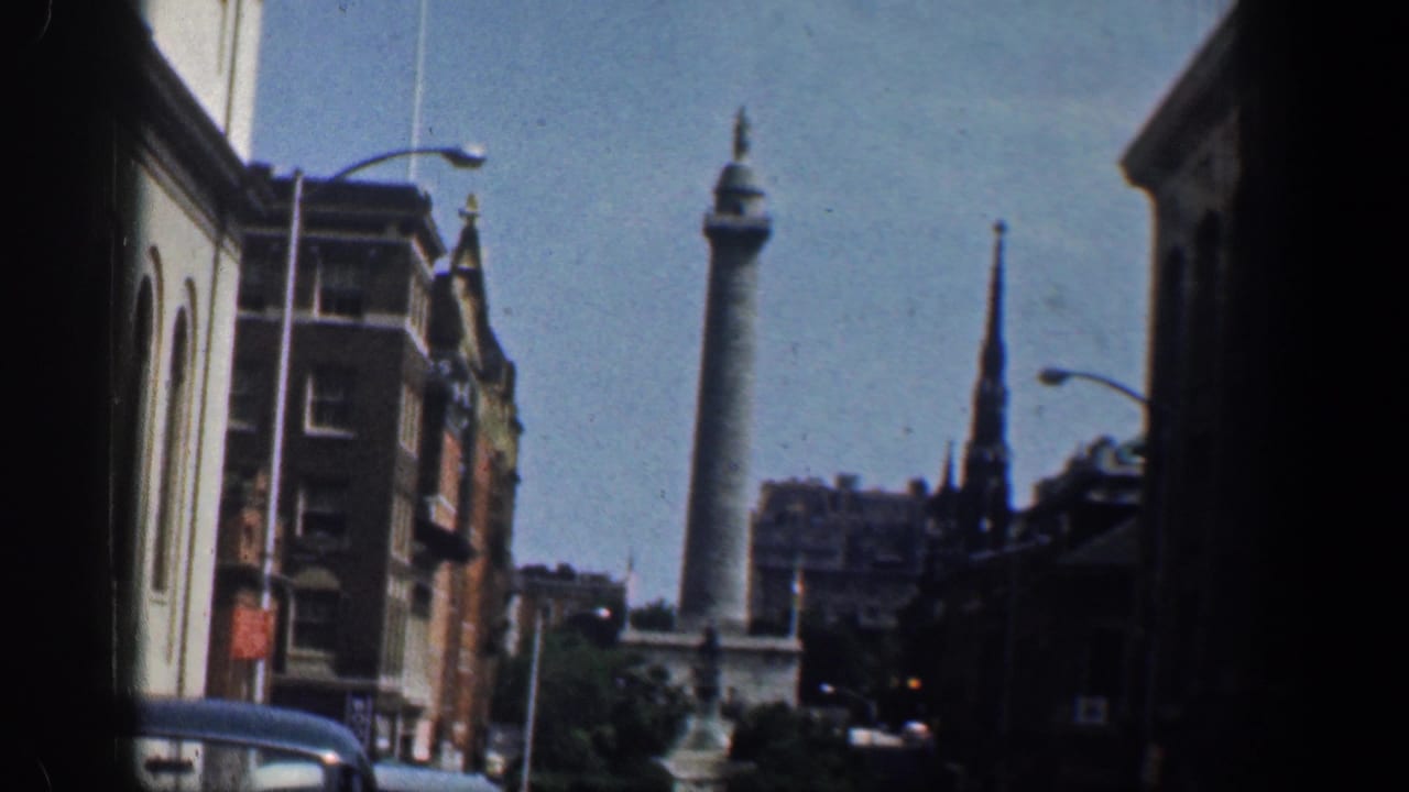 Street view with brick buildings, cars, streetlights, steeple, and a distant monument column.