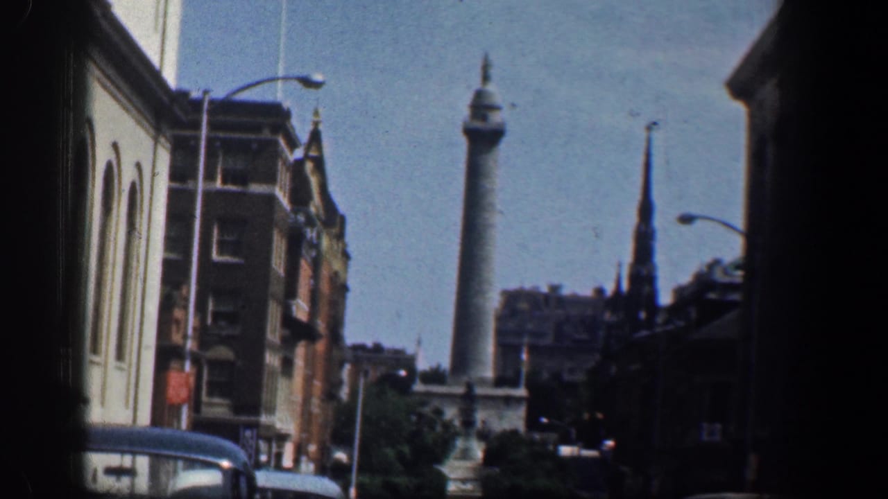 Washington DC street scene on 8mm film, with a distant civic column, a dark steeple, brick buildings, streetlights, and classic cars.