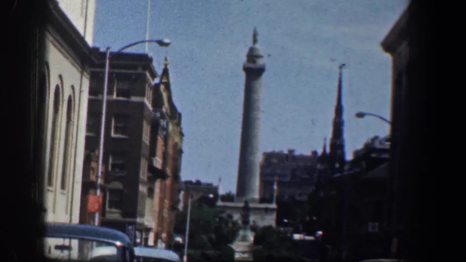 Washington DC street scene on 8mm film, with a distant civic column, a dark steeple, brick buildings, streetlights, and classic cars.