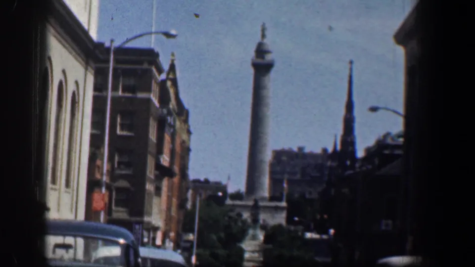 1961 Washington DC street footage shows classic automobiles below brick buildings, with a distant monument-like column and steeple against blue sky.