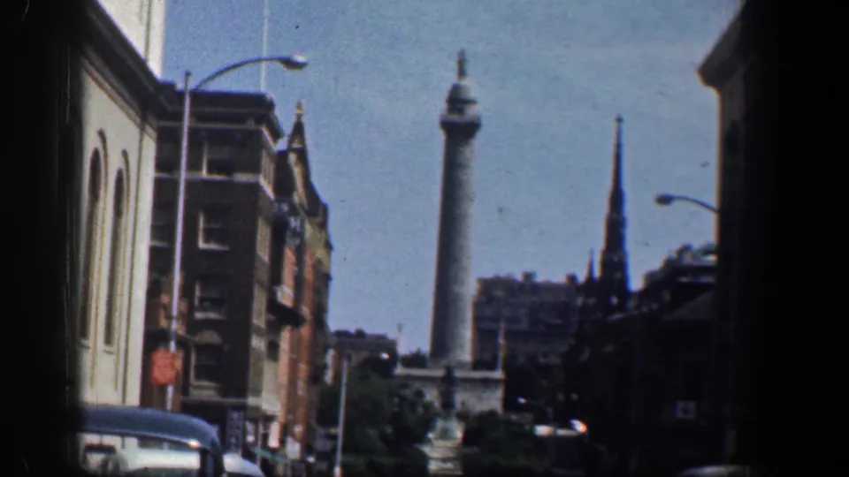 Washington DC home-movie frame with mid-century cars, brick facades, curved streetlights, a dark steeple, and a distant column-like monument.