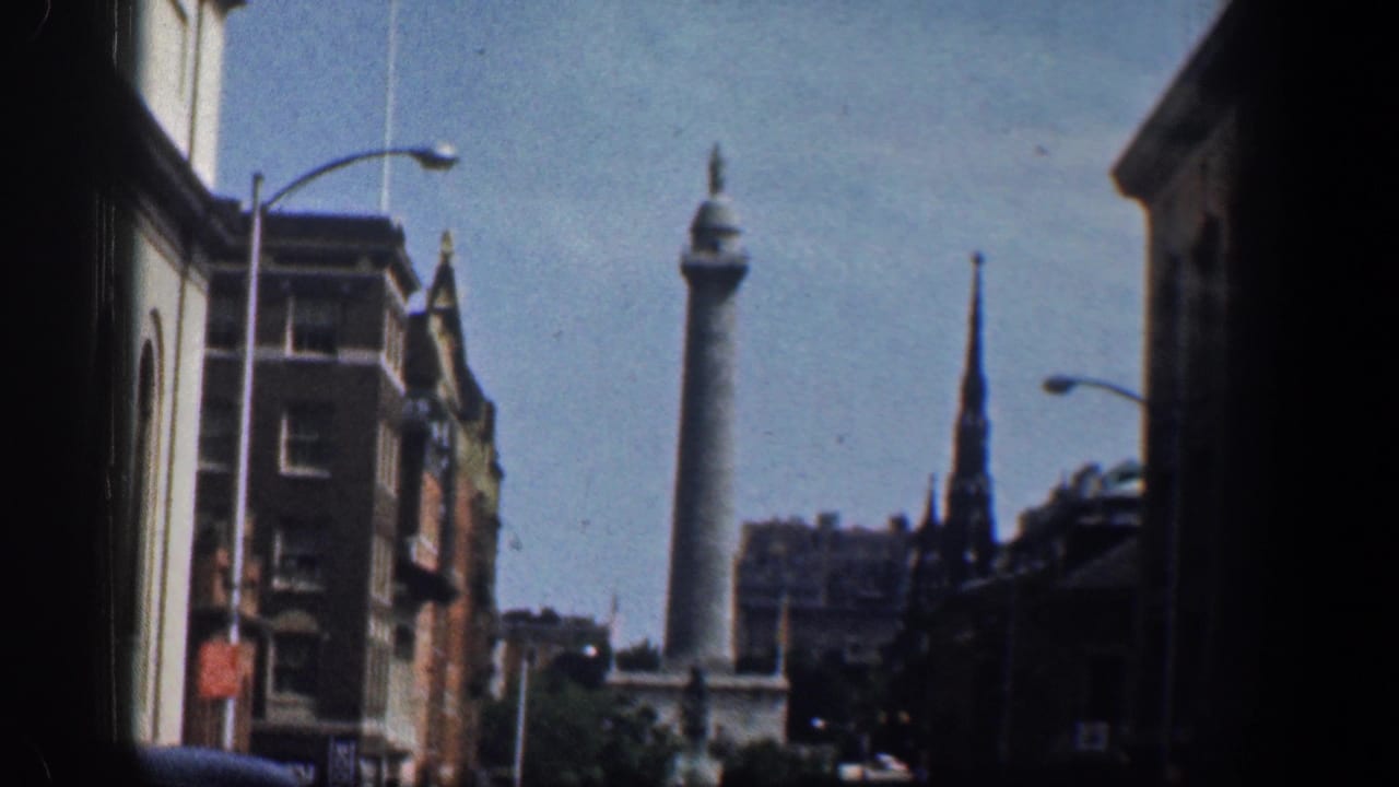 Street view with a tall stone monument, brick buildings, church spire, streetlights, and blue sky.