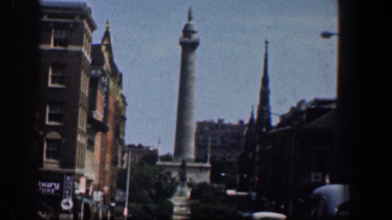 1961 Washington, DC street view with the Washington Monument beyond traffic, brick buildings, trees, and a church steeple.