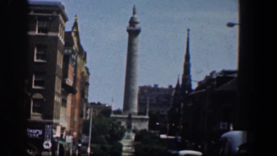 1961 Washington, DC street view with the Washington Monument beyond traffic, brick buildings, trees, and a church steeple.