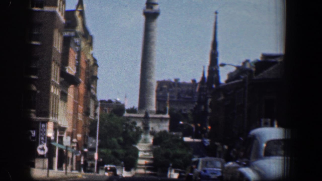 Washington Monument seen beyond downtown buildings, trees, traffic, and a church steeple.