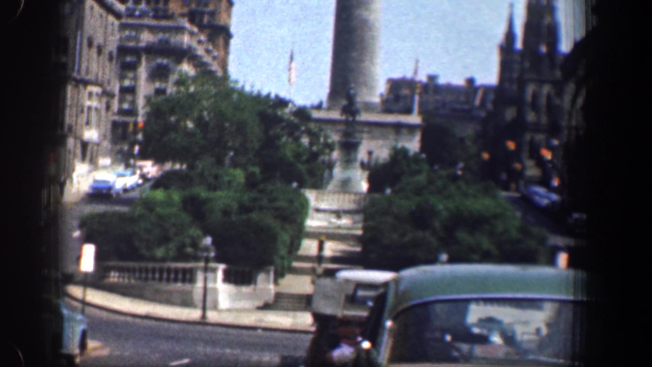 Green car in the foreground with a stone streetscape, trees, a tall column, and a distant Gothic spire.