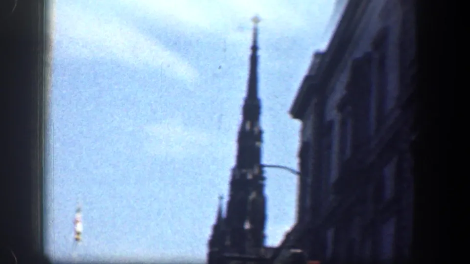 Handheld 1961 Washington DC view looking upward at a dark Gothic spire, blue sky, a building facade, and a curved streetlamp.