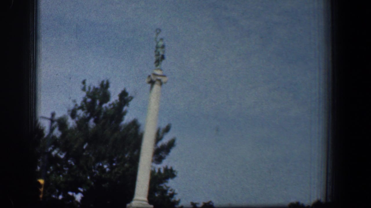1961 Washington, DC frame showing a white statue-topped column monument with trees below and blue sky behind it.