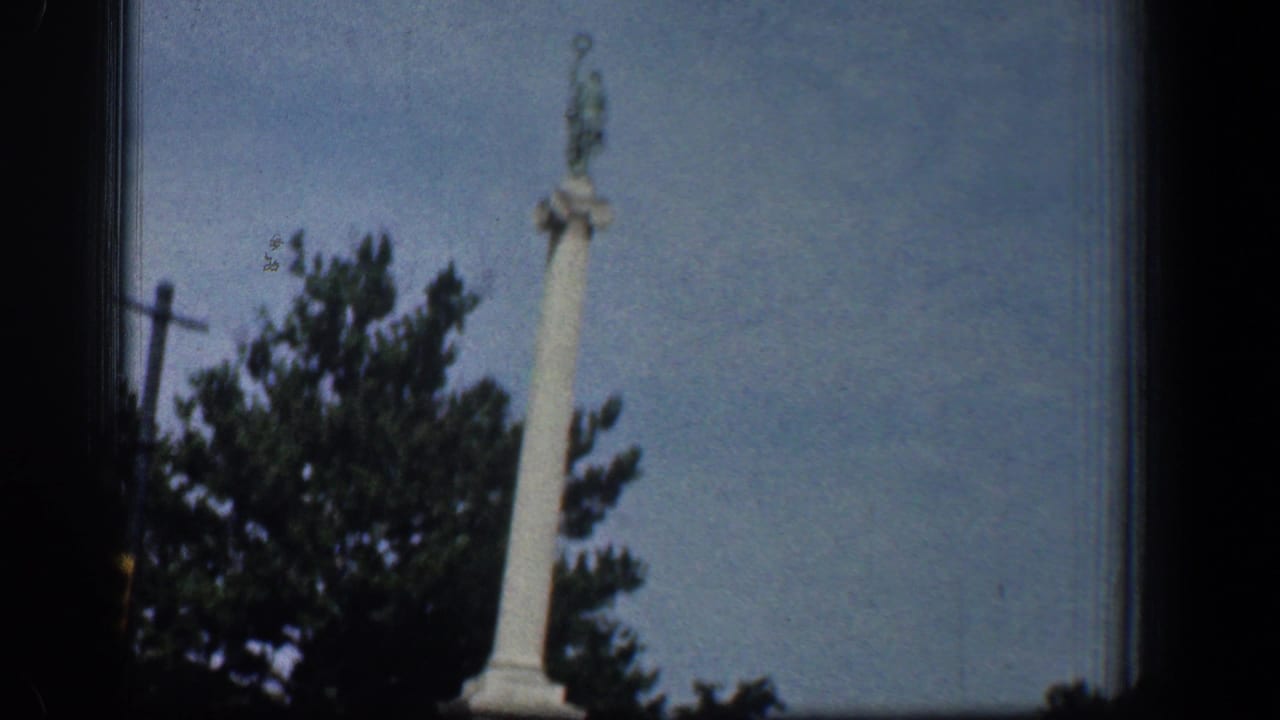 1961 Washington DC home-movie frame showing a tall white column monument with a statue top, partly obscured by trees from a moving car.