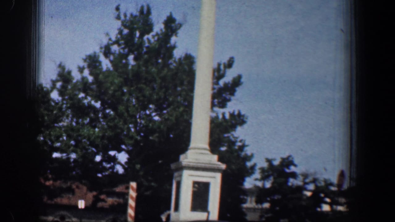 The white column monument is seen more fully from the road, with a square base, trees, and a striped sign in the Washington DC setting.