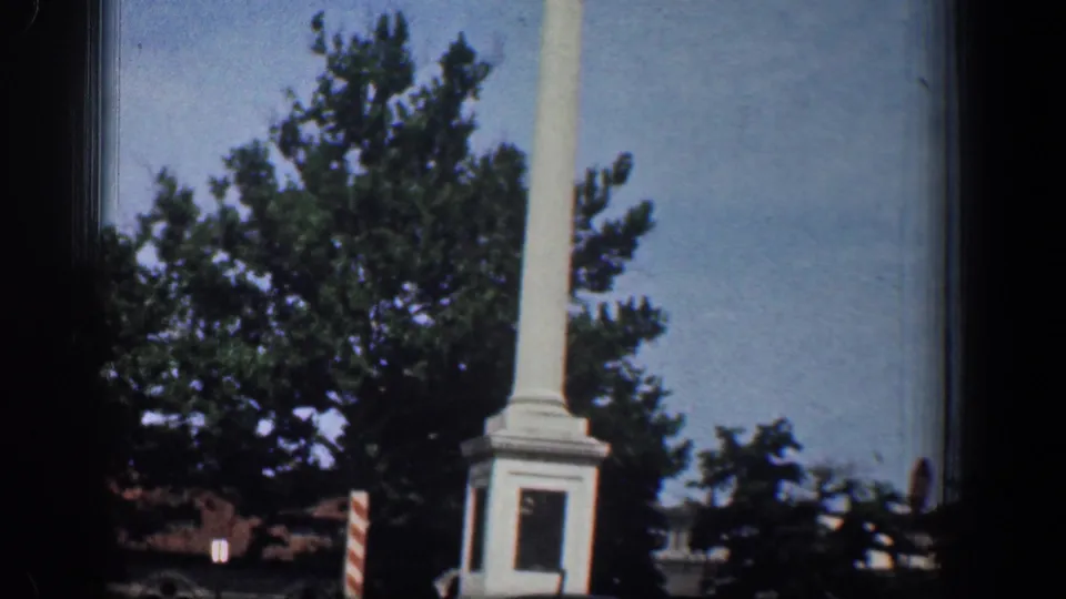 The white column monument is seen more fully from the road, with a square base, trees, and a striped sign in the Washington DC setting.