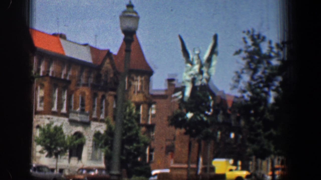 A winged statue stands before brick buildings with red roofs, with a streetlamp, trees, and cars visible in 1961 Washington DC footage.