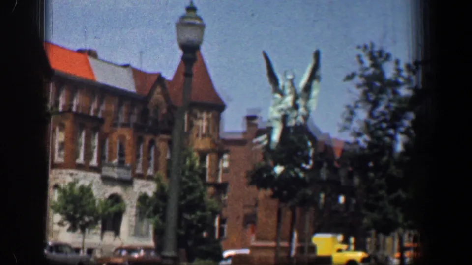 A winged statue stands before brick buildings with red roofs, with a streetlamp, trees, and cars visible in 1961 Washington DC footage.