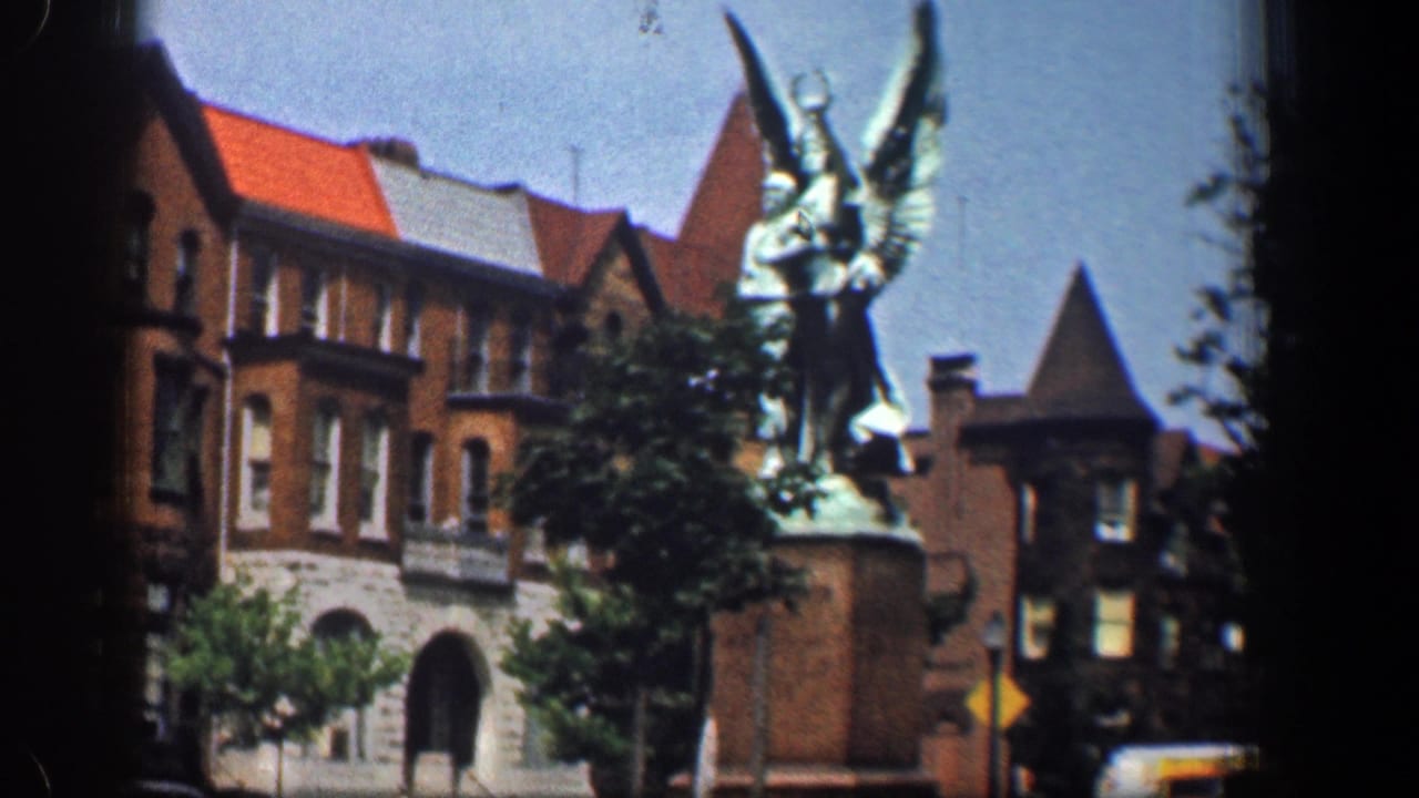 The winged statue on a pedestal is framed against historic-looking brick buildings, trees, and roadside traffic in Washington DC.