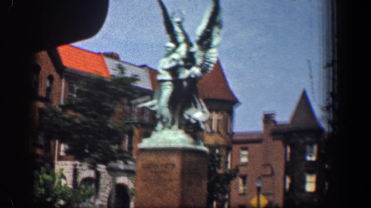 1961 Washington, DC home-movie frame showing a two-figure monument on a pedestal near brick buildings and trees from inside a moving vehicle.