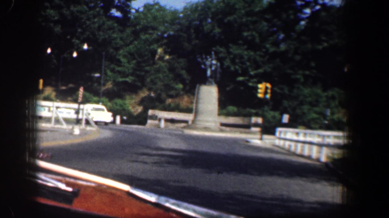 1961 Washington, DC street view from a moving vehicle approaching a monument near a traffic signal, white car, trees, and stone railing.