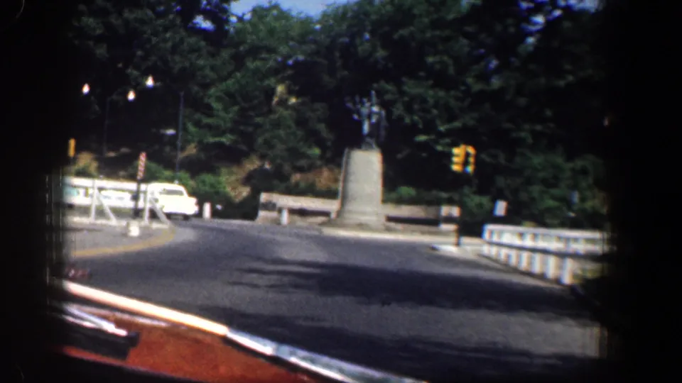 1961 Washington, DC street view from a moving vehicle approaching a monument near a traffic signal, white car, trees, and stone railing.