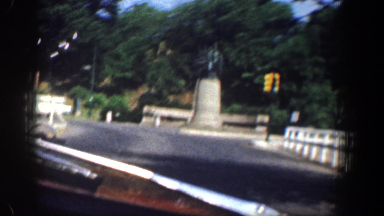 Motion-blurred 1961 Washington, DC frame showing the vehicle passing a traffic circle with a monument, white car, railing, and dense trees.