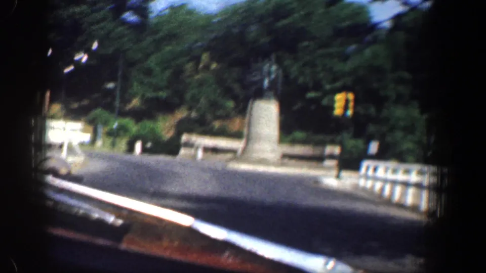 Motion-blurred 1961 Washington, DC frame showing the vehicle passing a traffic circle with a monument, white car, railing, and dense trees.