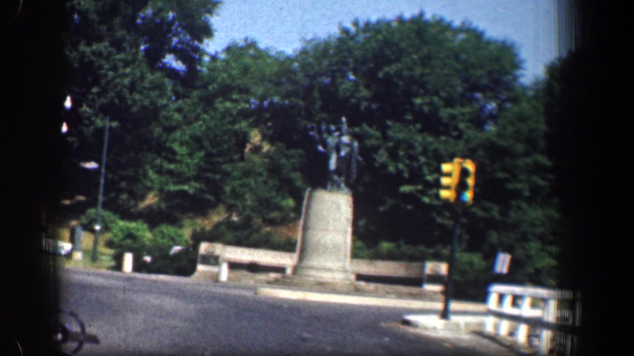 Traffic circle with a statue on a stone pedestal, benches, trees, and a yellow signal light.