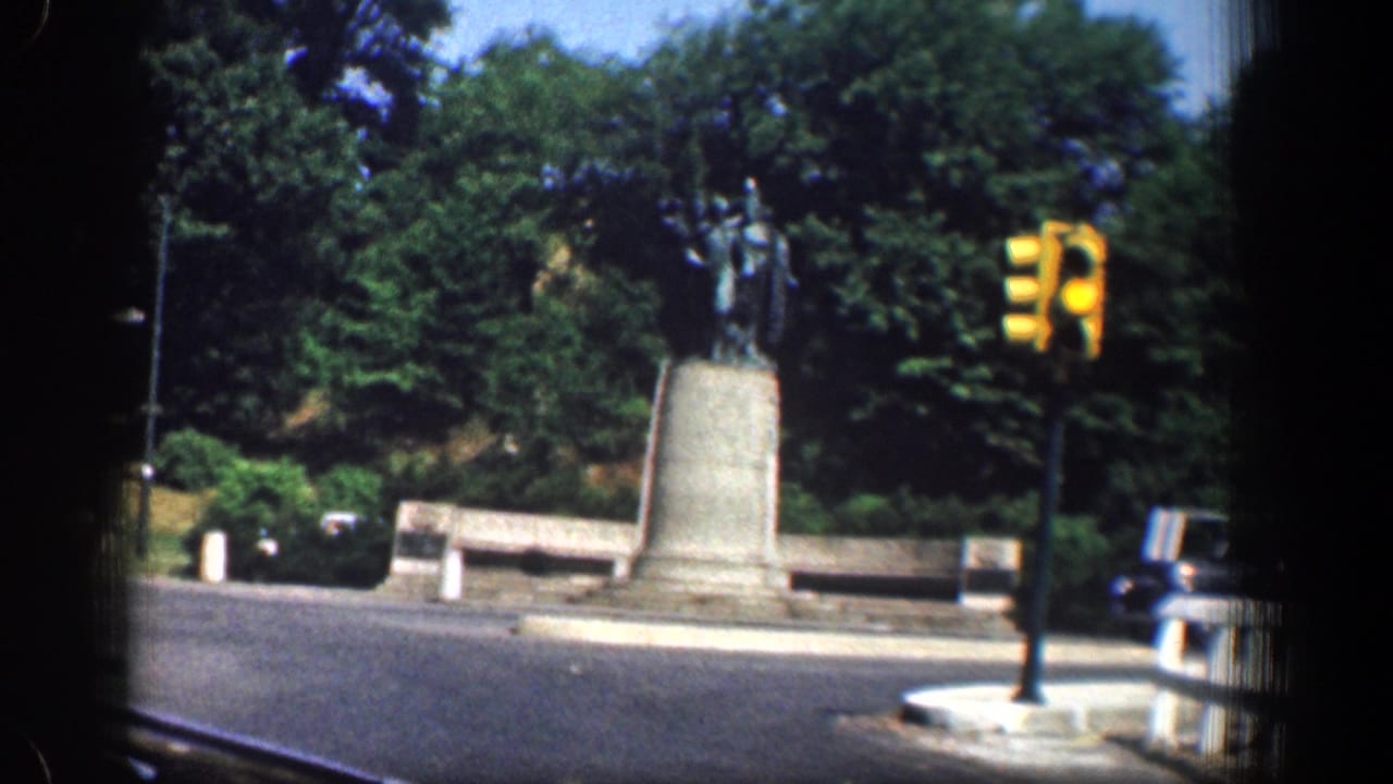 1961 Washington, DC frame of a statue at a landscaped traffic circle, with benches, trees, street pavement, and a yellow traffic signal visible.