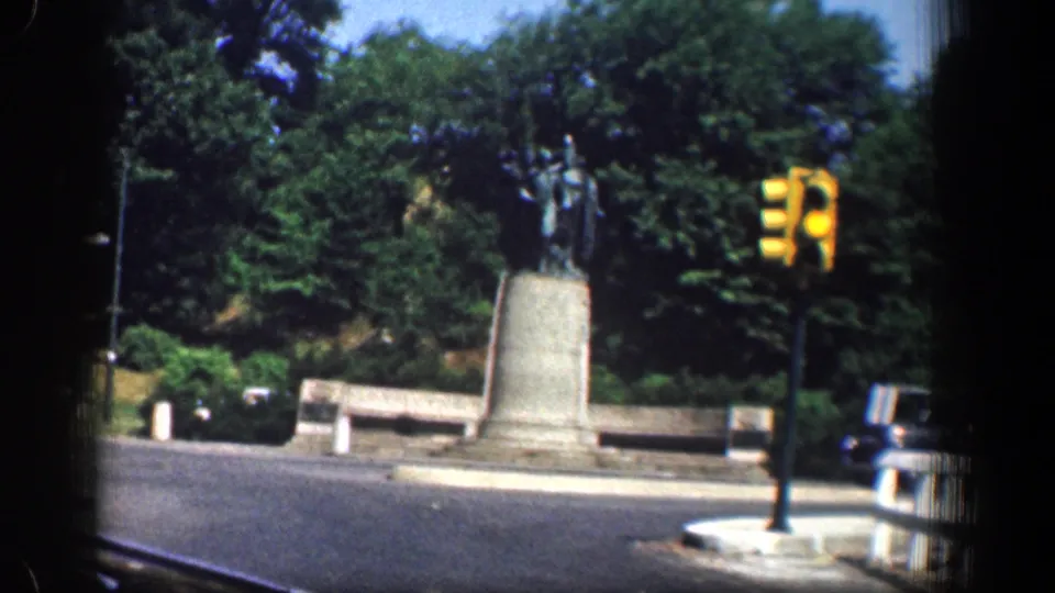 1961 Washington, DC frame of a statue at a landscaped traffic circle, with benches, trees, street pavement, and a yellow traffic signal visible.