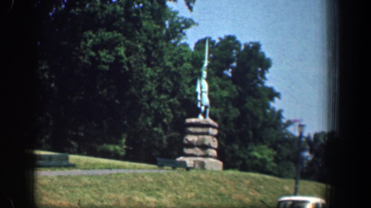 1961 Washington, DC home-movie view of a statue on a stone base above grass, framed by trees with a lamppost and street edge in view.