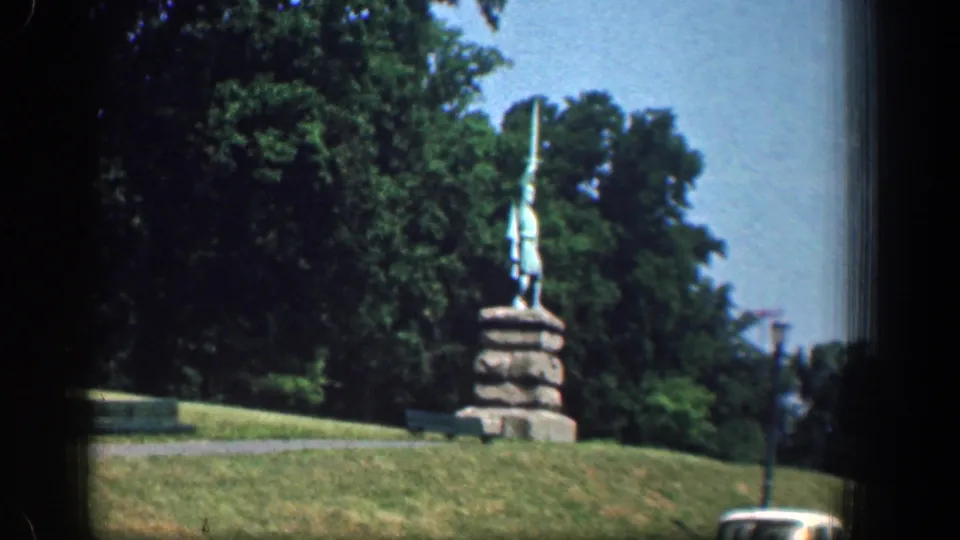 1961 Washington, DC home-movie view of a statue on a stone base above grass, framed by trees with a lamppost and street edge in view.