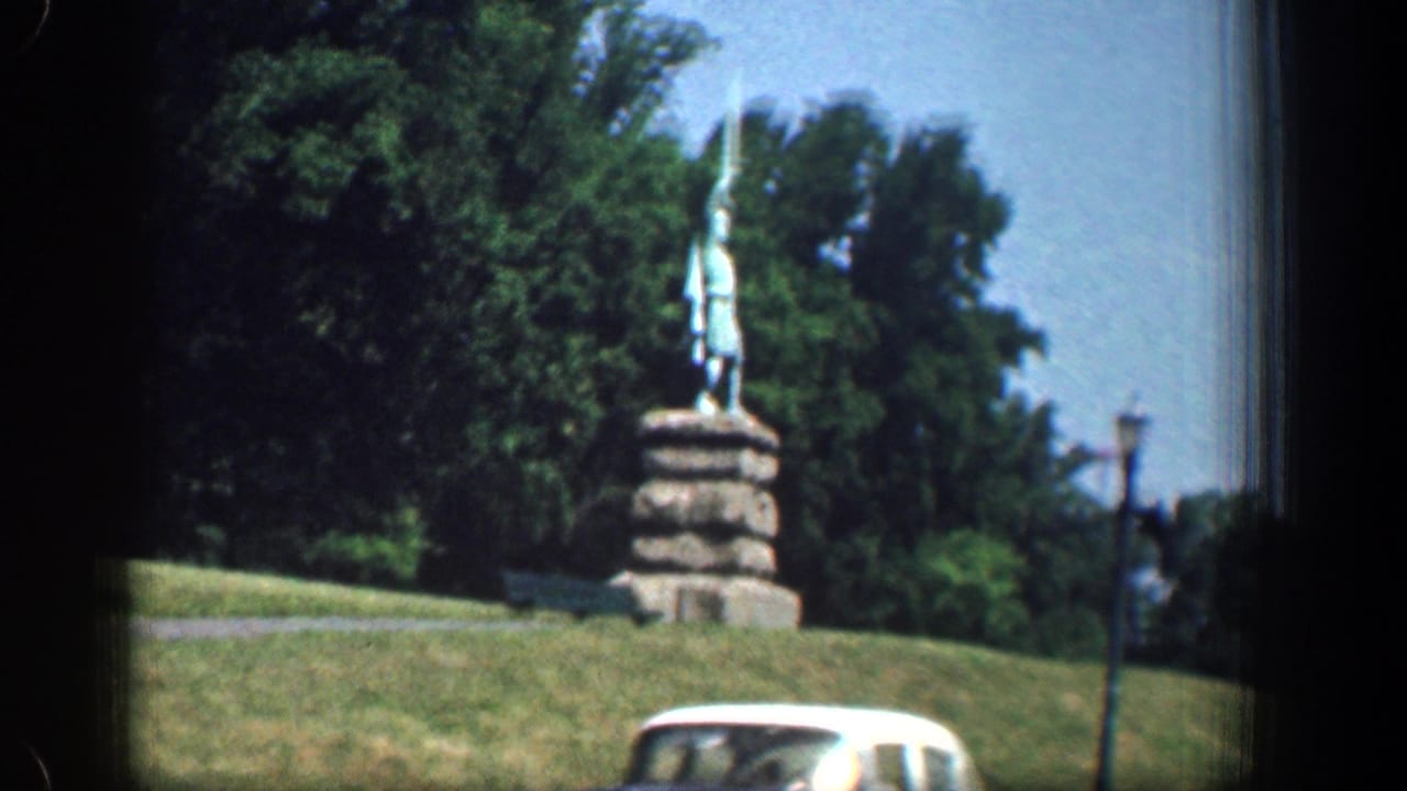 1961 Washington, DC frame showing a public statue on a stone pedestal above a grassy slope, with trees, a lamppost, and a white car passing below.