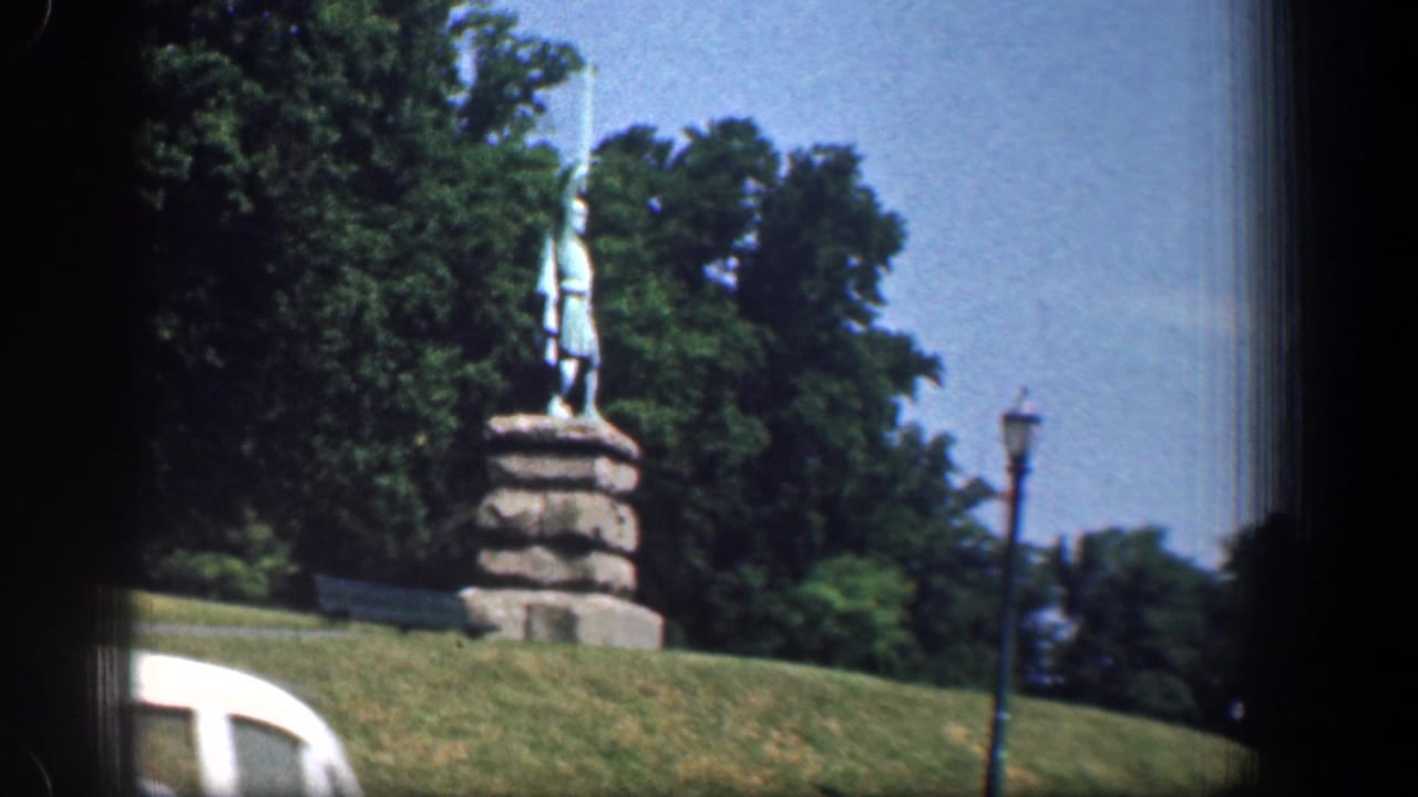 1961 Washington DC home-movie frame showing a statue on a stone pedestal, green lawn, trees, a roadside lamp, and part of a white car.
