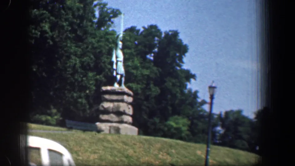 1961 Washington DC home-movie frame showing a statue on a stone pedestal, green lawn, trees, a roadside lamp, and part of a white car.