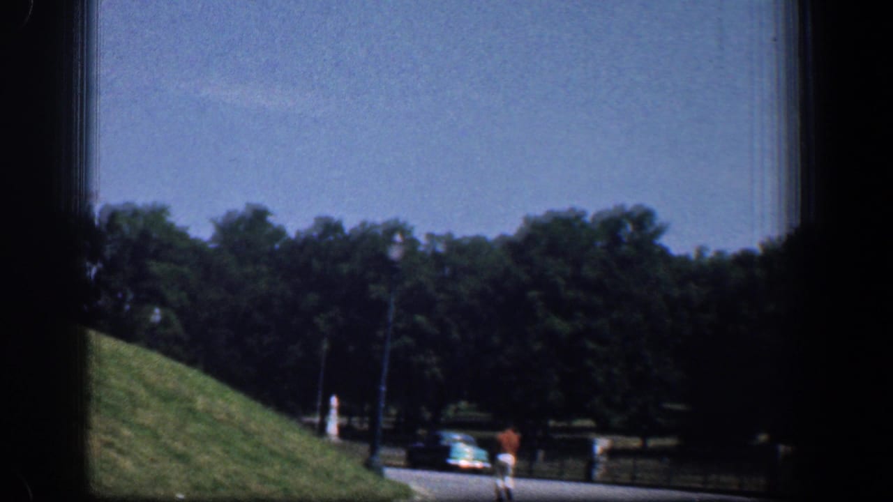 1961 Washington DC frame showing a landscaped roadside with trees, a blue classic car, a darker vehicle, and distant pedestrians near the park edge.