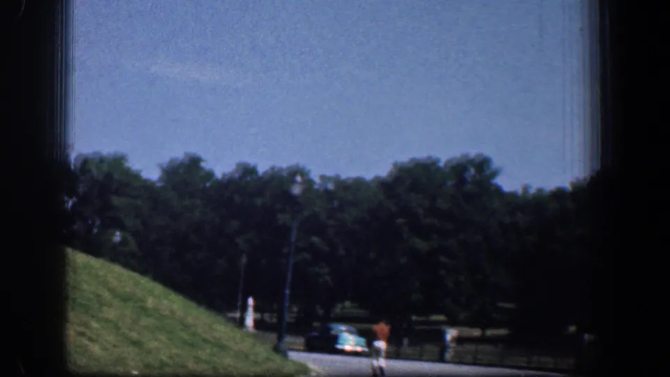 1961 Washington DC frame showing a landscaped roadside with trees, a blue classic car, a darker vehicle, and distant pedestrians near the park edge.