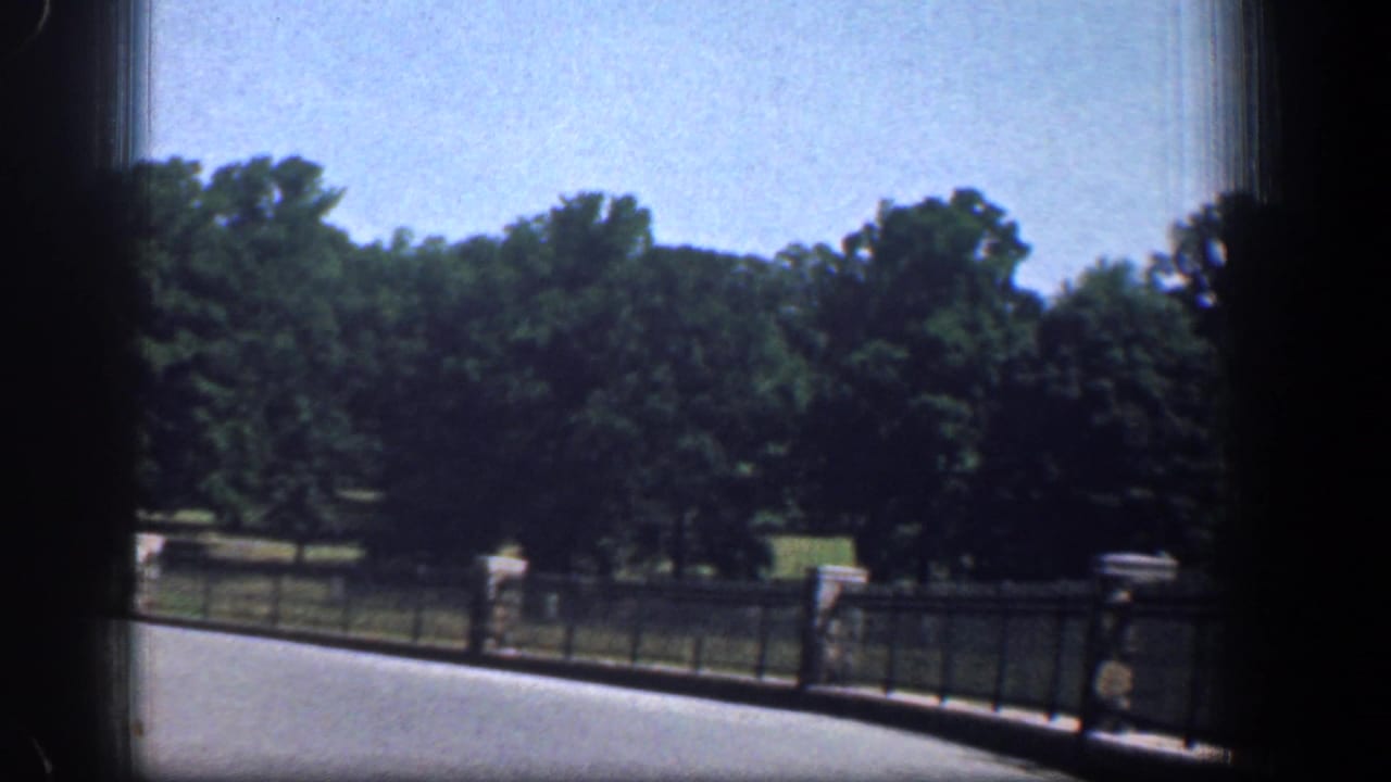 1961 Washington DC park view from a vehicle, with black fencing, stone posts, green trees, roadway, and a distant walking person.