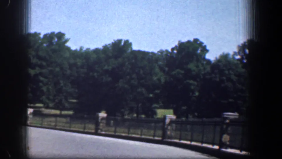 1961 Washington DC park view from a vehicle, with black fencing, stone posts, green trees, roadway, and a distant walking person.
