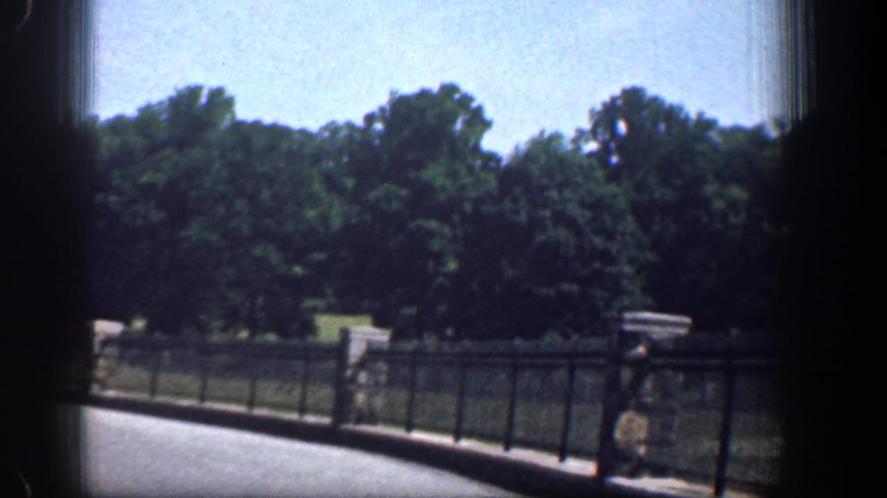 Vehicle-borne 1961 Washington DC park footage showing a black fence, stone posts, shaded trees, grass, and a distant pedestrian.
