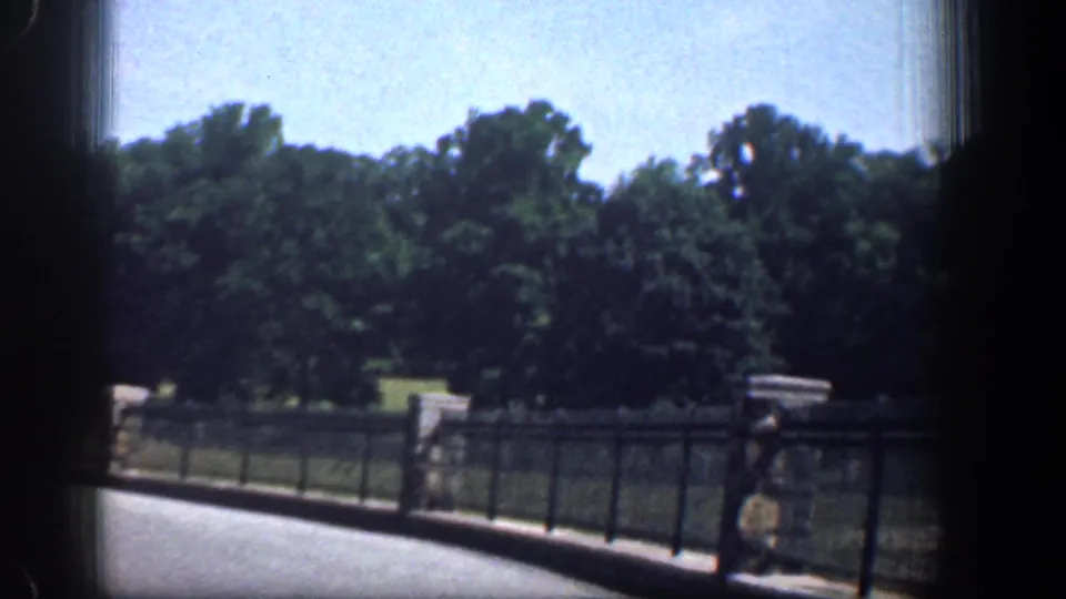 Vehicle-borne 1961 Washington DC park footage showing a black fence, stone posts, shaded trees, grass, and a distant pedestrian.