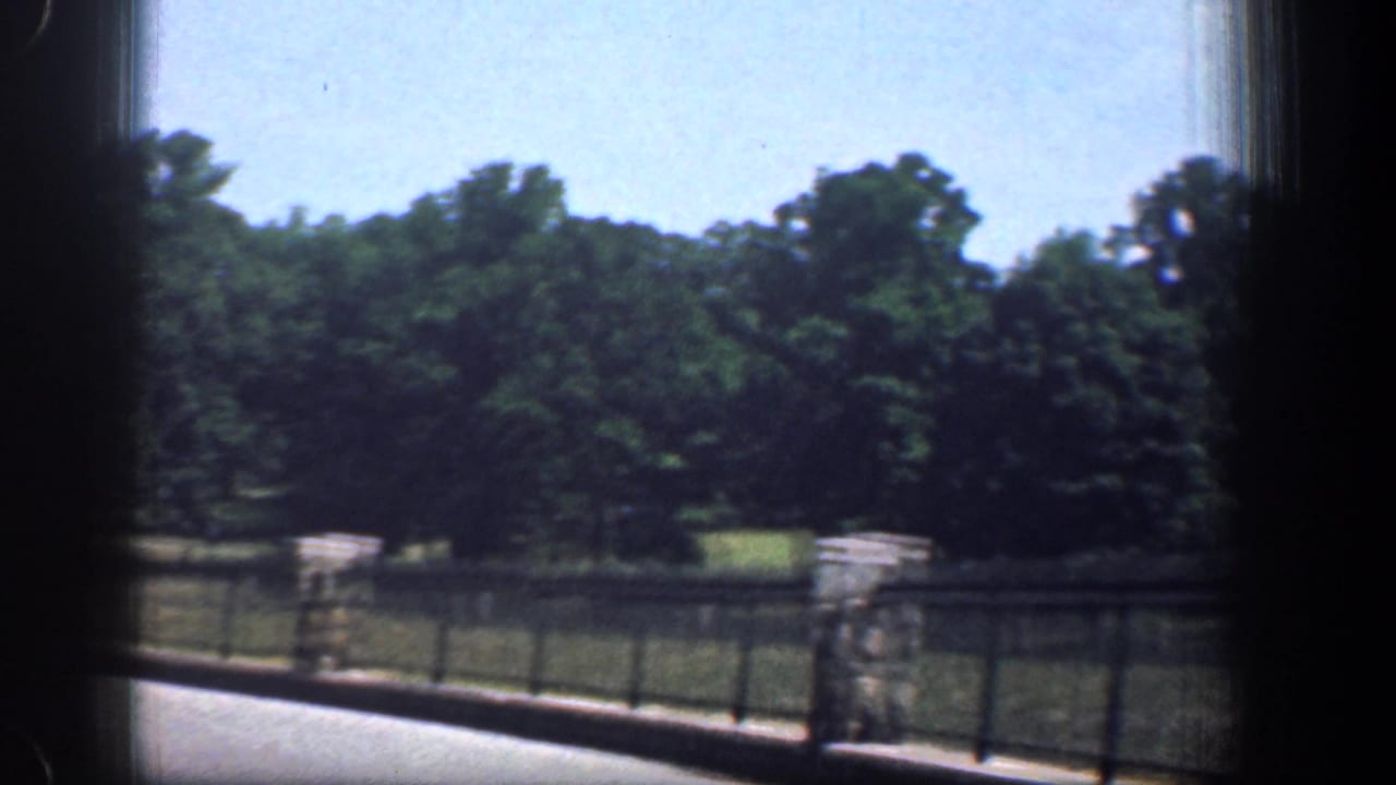 Late-frame 1961 Washington DC park view with trees, grass, black railing, stone posts, road edge, and dark car-window borders.