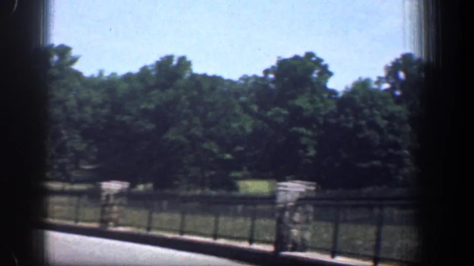 Late-frame 1961 Washington DC park view with trees, grass, black railing, stone posts, road edge, and dark car-window borders.