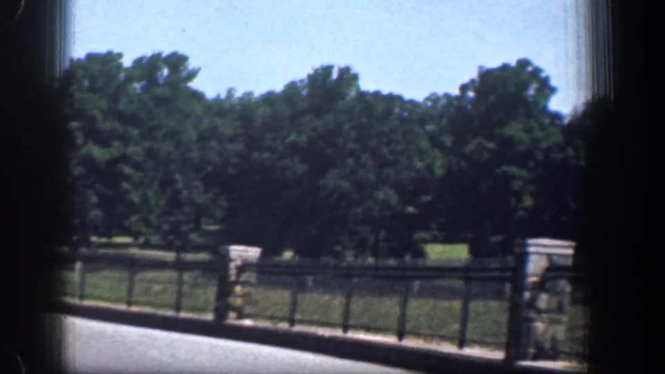 1961 Washington, DC home-movie view of a road, stone gatepost, metal fence, grass, and trees seen through a vehicle window.