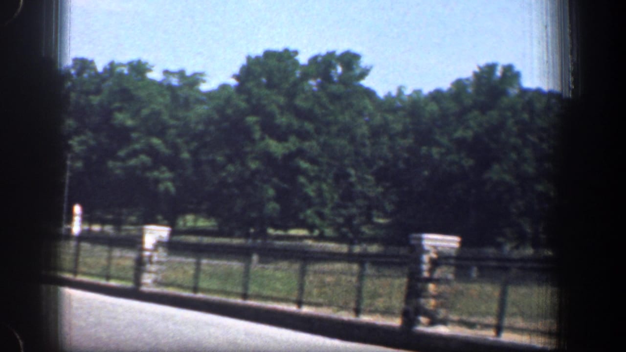 Washington, DC road scene from 1961 with metal fencing, stone pillars, grass, trees, and dark window framing from the vehicle.