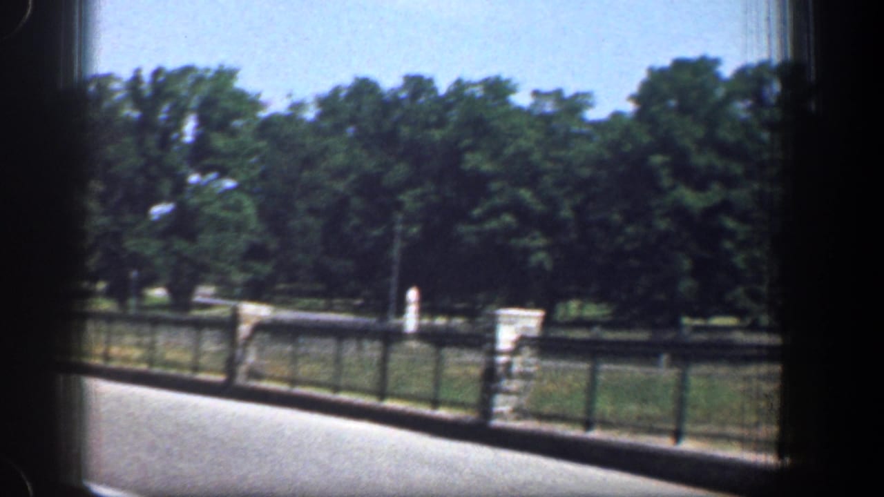 1961 Washington, DC car-window view showing a fenced roadside, stone post, grass, trees, and open sky along the roadway.