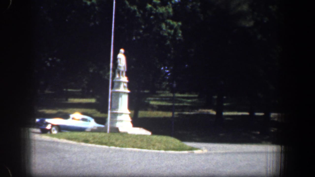 1961 Washington, DC roadside view with a statue on a grassy median, a light-colored vintage car, a pole, road, and shaded trees.