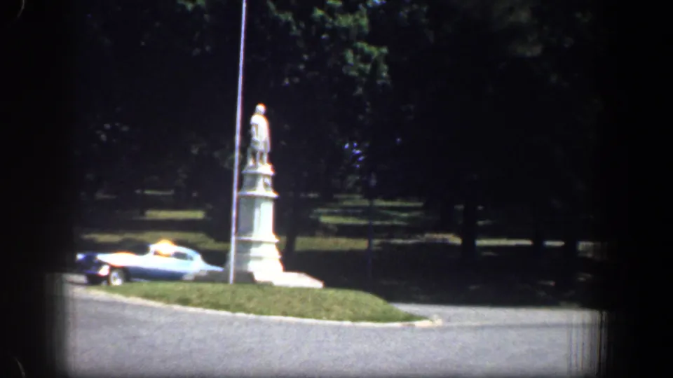 1961 Washington, DC roadside view with a statue on a grassy median, a light-colored vintage car, a pole, road, and shaded trees.