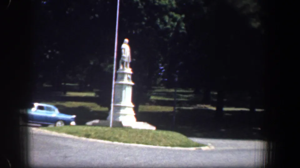 1961 Washington, DC home-movie frame showing a white stone statue beside a road, a blue sedan, grass, and trees.