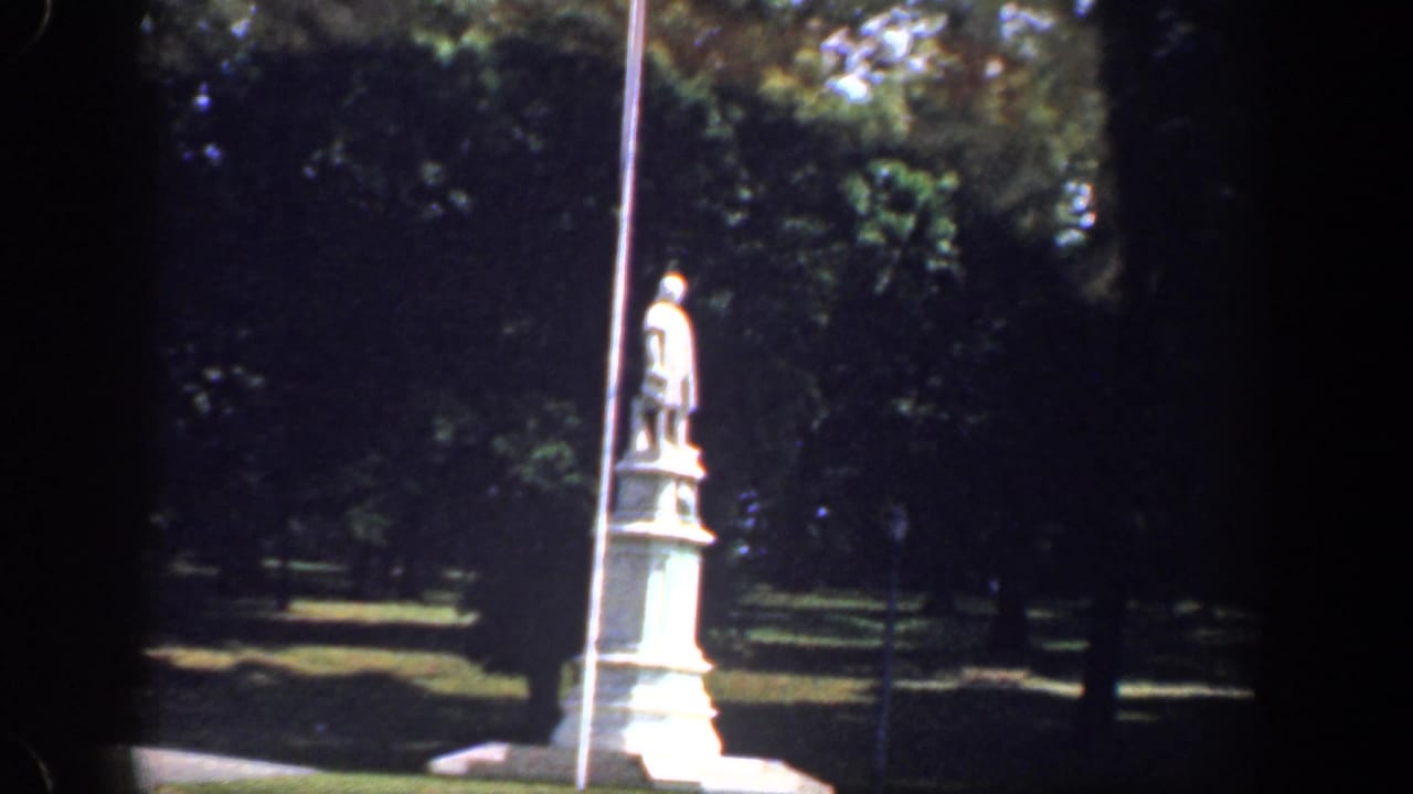 1961 Washington, DC frame with a roadside statue, tall pole, paved curve, and shaded trees in the background.