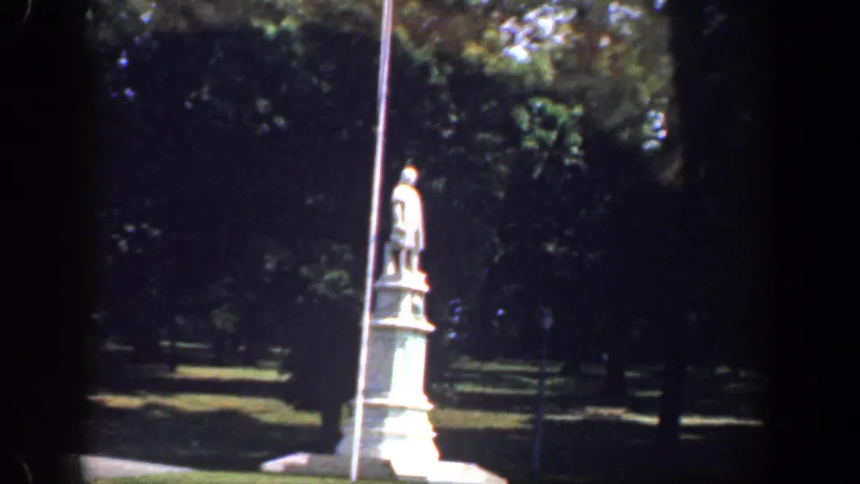 1961 Washington, DC frame with a roadside statue, tall pole, paved curve, and shaded trees in the background.