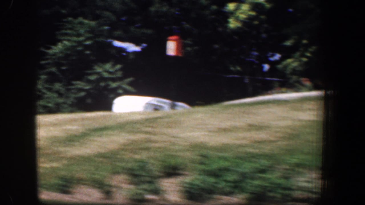 1961 8mm drive-by frame with lawn, low plantings, shaded trees, and a pale vehicle or trailer shape partly visible.