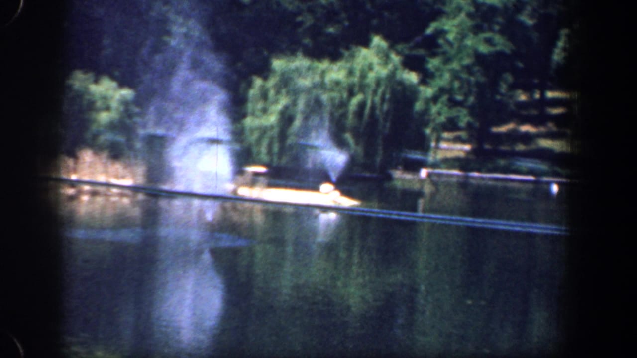 Home-movie frame from 1961 Washington, DC showing fountain spray across a park lake near a willow and sunlit shore.