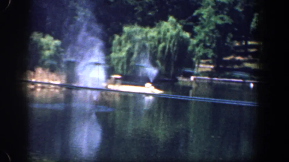 Home-movie frame from 1961 Washington, DC showing fountain spray across a park lake near a willow and sunlit shore.